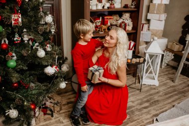 Kırmızı elbiseli mutlu anne ve oğlu, Noel hediyesini süslenmiş bir ağacın ve tatil raflarının yanında paylaşıyorlar, sıcak bir aile bağı kurma anını yakalıyorlar.