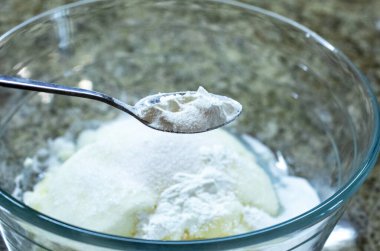 Beyaz un ya da nişasta tutan gümüş kaşığı yakın plan, bir mutfakta süzme peynir ve şeker de dahil olmak üzere cam bir kase fırın malzemesine ekliyor.