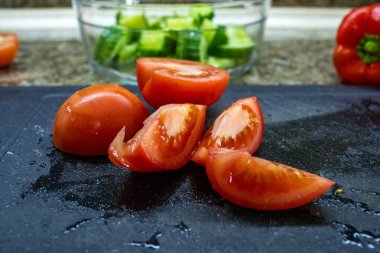 Kırmızı biber dilimlenmiş kırmızı domateslerin yakın plan fotoğrafı. Mutfak arka planında bir salata hazırlarken kırmızı biber ve dilimlenmiş bir kase salatalık.