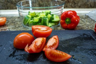 Kırmızı biber dilimlenmiş kırmızı domateslerin yakın plan fotoğrafı. Mutfak arka planında bir salata hazırlarken kırmızı biber ve dilimlenmiş bir kase salatalık.