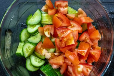 Taze doğranmış domatesler ve dilimlenmiş salatalıklarla dolu temiz bir cam kasenin tepesinde sağlıklı, çiğ sebze salatasının canlı kırmızı ve yeşil renklerini gösteriyor.