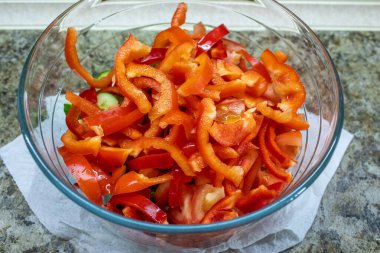 Taze dilimlenmiş kırmızı çan biberleri, domatesler ve salatalıkla dolu, sağlıklı, renkli bir sebze salatası gibi giyinmeye hazır, parlak bir cam kasenin yukarıdan aşağı manzarası.