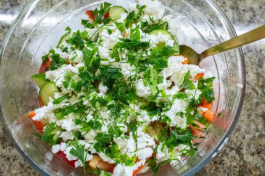 Taze sebze salatasıyla dolu büyük ve berrak bir cam kasenin tepesinde beyaz peynirli feta ya da kır evi ve doğranmış yeşil otların karışımı var.