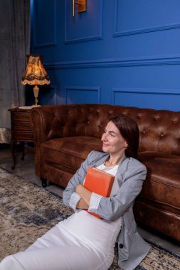 Bir kitapla yerde oturan rahat kadın, lüks mavi ve kahverengi bir iç mekanda eski bir deri kanepeye yaslanıyor.