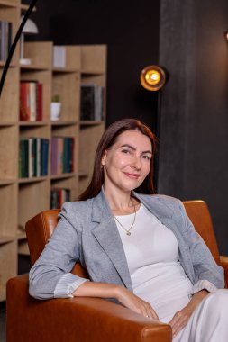 Zarif bir iş kadını modern bir deri koltukta oturuyor ve şık, karanlık bir ofiste, arka planda bir kitaplığa karşı poz veriyor.