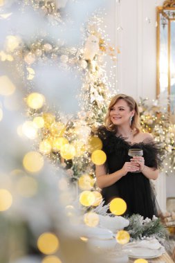 Siyah tüllü elbise ve elmas takılar içinde zarif bir kadın Noel ve yeni yılı kutluyor. Bir bardak şampanyayla ışıl ışıl Noel ağaçlarının arka planında.