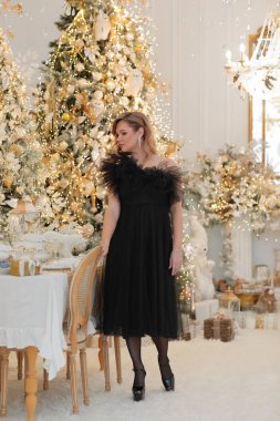 Siyah tüllü elbisesi ve yüksek topuklu ayakkabıları olan zarif bir kadın bir sandalyenin yanında duruyor, lüks bir altın ve beyaz bir Noel 'de poz veriyor ve yeni yıl dekorasyonu yapıyor.