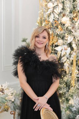 Siyah tüllü elbise ve elmas takılar içinde zarif bir kadın Noel ve yeni yılı kutluyor. Bir bardak şampanyayla ışıl ışıl Noel ağaçlarının arka planında.