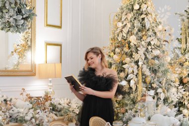 Siyah tül elbise ve mücevher takmış zarif bir kadın lüks bir altın ve beyaz Noel 'de bir kitap ya da resim çerçevesine ve dekore edilmiş yeni yıla bakıyor.