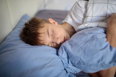 Mavi bir yastıkta mışıl mışıl uyuyan, yatağında yorganla rahatça uyuyan barışçıl bir gencin yakın çekimi.