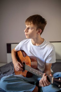 Yaratıcı genç çocuk yatağında oturmuş akustik gitar çalıyor ve not defterine şarkı sözleri ya da notlar yazıyor.