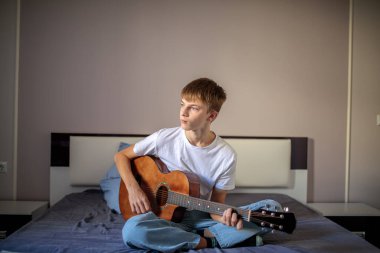 Yaratıcı genç çocuk yatağında oturmuş akustik gitar çalıyor ve not defterine şarkı sözleri ya da notlar yazıyor.