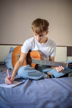 Yaratıcı genç çocuk yatağında oturmuş akustik gitar çalıyor ve not defterine şarkı sözleri ya da notlar yazıyor.