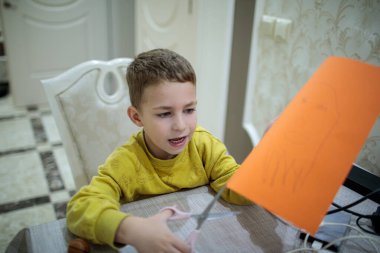 Masada oturan sevimli küçük çocuk okul projesi için portakal renkli kağıtları makasla kesiyor.