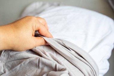 Yapılmamış bir yatakta, kumaş dokusuna odaklanmış nötr renkli çarşafları çeken ya da düzenleyen bir elin yakın çekimi.