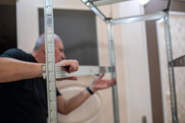 Orta yaşlı bir adam, parlak döşemeleri ve dekoratif duvar kağıtları olan zarif bir odada galvanize metal bir raf ünitesi kuruyor.