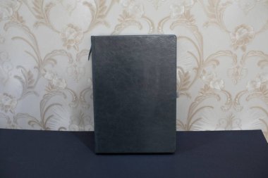 Dikey siyah deri ya da sahte deri kaplı defter koyu bir yüzey üzerinde eski moda duvar kâğıdının arka planında duruyor.