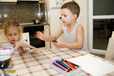 Mutfak masasında el işi yapan iki küçük kardeş, kağıt kesmeye ve renkli kalemlerle çalışmaya konsantre oluyorlar.
