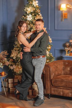 Romantik, şık bir çift, Noel arifesini ya da Noel arifesini kutlayan, lüks, koyu mavi bir iç mekanda altın madalyalı bir Noel ağacının yanında poz veriyorlar.