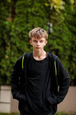 Ciddi bir gencin portresi ya da genç bir çocuğun elleri kapüşonlu cebinde dışarıda dikilirken bulanık yeşil çit arka planına karşı günlük siyah kıyafetler giymesi.