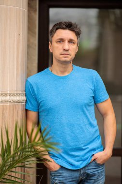 Mavi tişörtlü ve kot pantolonlu sakin orta yaşlı bir adamın portresi. Açık havada bir sütuna yaslanmış ve elleri ceplerinde kameraya bakıyor.