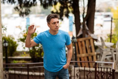 Mavi tişörtlü ve kot pantolonlu orta yaşlı bir adamın portresi, dışarıda duruyor ve elini kaldırıyor. Tarafsız ya da ciddi bir ifadeyle selam vermek ya da durdurmak için.