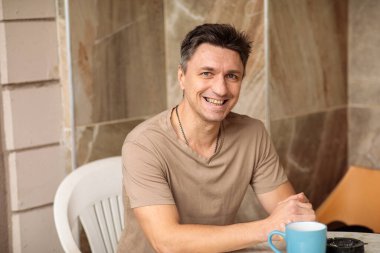 Orta yaşlı, mutlu, mutlu, orta yaşlı bir adamın açık hava avlusunda kahve fincanıyla oturduğu yakın plan portresi.