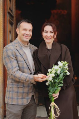 Kameraya gülümseyen mutlu, zarif bir çift. Karanlık, tarihi bir girişin önünde el ele tutuşup bir buket beyaz çiçek.