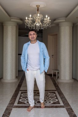 Mutlu, orta yaşlı, çıplak ayaklı bir adam. Sütunları ve avizesi olan zarif bir koridorda duruyor. Elleri ceplerinde.