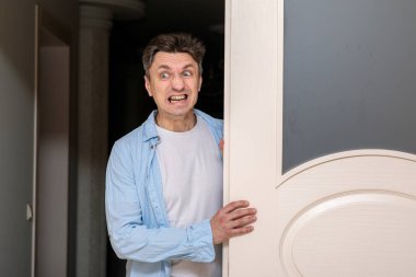 Orta yaşlı bir adam öfkeli, kızgın ya da şok olmuş bir ifadeyle aralık kapısından gizlice bakıyor, dişlerini sıkıyor.