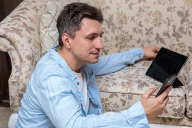 Orta yaşlı bir adam yerde oturuyor, akıllı telefonu ve tabletine odaklanmış, lüks bir kanepeye karşı çalışıyor ya da dinleniyor.