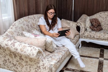 Gözlüklü ve günlük beyaz elbiseli genç bir kadın lüks bir çiçek kanepesinde dinleniyor, dijital tablet okumaya ya da onunla çalışmaya odaklanıyor.