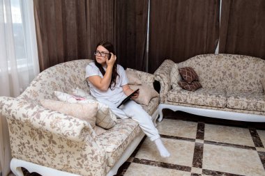 Gözlüklü ve günlük beyaz elbiseli genç bir kadın lüks bir çiçek kanepesinde dinleniyor, dijital tablet okumaya ya da onunla çalışmaya odaklanıyor.