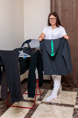 Beyaz giysili genç bir kadın mutlu bir şekilde asılı ya da katlanmış koyu renkli çamaşırları kırmızı bir askıda seramik bir zeminde.