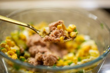 Ev yapımı sağlıklı bir salata hazırlarken cam kaseden ton balığı, mısır ve yumurta karışımını kaldıran altın kaşığı yakın plan.