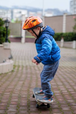 Sıcak mavi ceketli ve kasklı genç bir çocuk sonbaharda tuğladan bir kaldırımda kaykay sürmeyi öğreniyor.