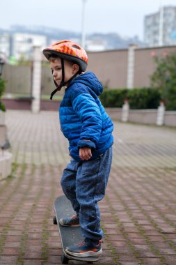 Sıcak mavi ceketli ve kasklı genç bir çocuk sonbaharda tuğladan bir kaldırımda kaykay sürmeyi öğreniyor.