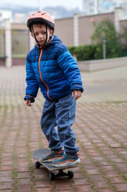 Sıcak mavi ceketli ve kasklı genç bir çocuk sonbaharda tuğladan bir kaldırımda kaykay sürmeyi öğreniyor.