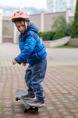 Sıcak mavi ceketli ve kasklı genç bir çocuk sonbaharda tuğladan bir kaldırımda kaykay sürmeyi öğreniyor.