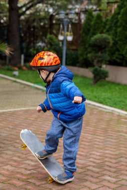 Sıcak mavi ceketli ve kasklı genç bir çocuk sonbaharda tuğladan bir kaldırımda kaykay sürmeyi öğreniyor.