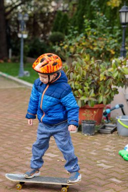 Sıcak mavi ceketli ve kasklı genç bir çocuk sonbaharda tuğladan bir kaldırımda kaykay sürmeyi öğreniyor.