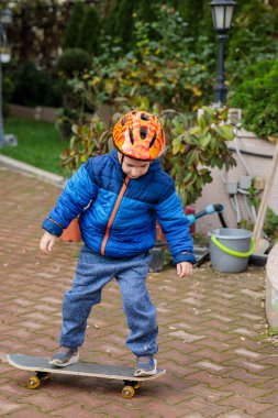 Sıcak mavi ceketli ve kasklı genç bir çocuk sonbaharda tuğladan bir kaldırımda kaykay sürmeyi öğreniyor.