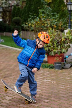 Sıcak mavi ceketli ve kasklı genç bir çocuk sonbaharda tuğladan bir kaldırımda kaykay sürmeyi öğreniyor.