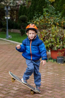 Sıcak mavi ceketli ve kasklı genç bir çocuk sonbaharda tuğladan bir kaldırımda kaykay sürmeyi öğreniyor.