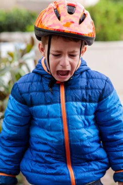 Emniyet kaskı takmış üzgün genç bir çocuk, düştükten sonra yoğun bir şekilde ağlıyor ya da dışarıda oynarken hayal kırıklığı yaşıyor.