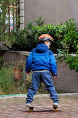 Sıcak mavi ceketli ve kasklı genç bir çocuk sonbaharda tuğladan bir kaldırımda kaykay sürmeyi öğreniyor.