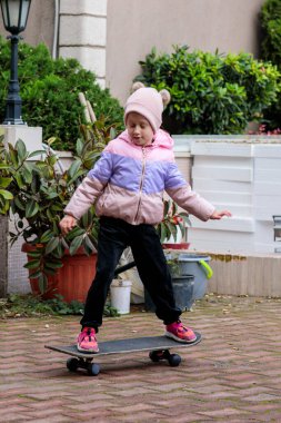 Renkli ceketli ve örgü şapkalı şirin küçük kız kaykaylı terasta diğer oyuncaklarla dengeyi kurmayı öğreniyor.