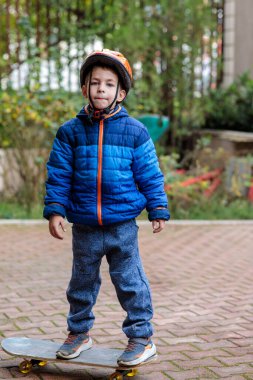 Sıcak mavi ceketli ve kasklı genç bir çocuk sonbaharda tuğladan bir kaldırımda kaykay sürmeyi öğreniyor.