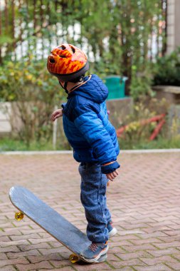 Sıcak mavi ceketli ve kasklı genç bir çocuk sonbaharda tuğladan bir kaldırımda kaykay sürmeyi öğreniyor.