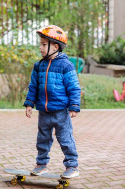 Sıcak mavi ceketli ve kasklı genç bir çocuk sonbaharda tuğladan bir kaldırımda kaykay sürmeyi öğreniyor.